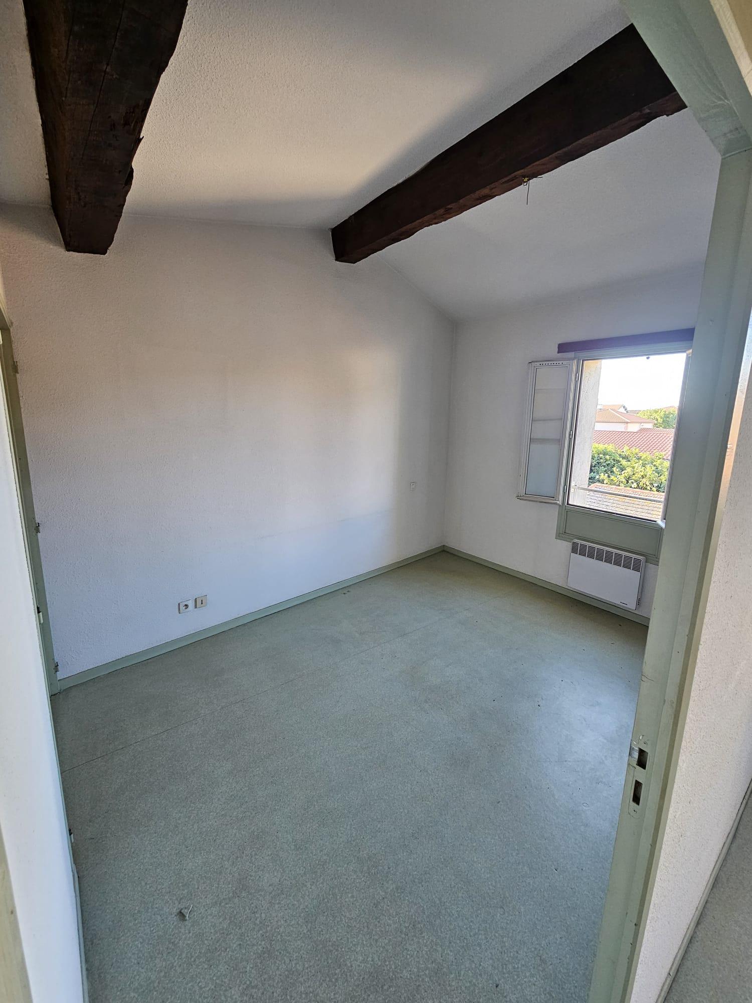 Chambre sous combles avant travaux, poutres sombres, sol souple vert, fenêtre à volets